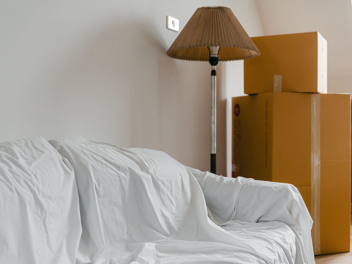 A sheet-covered sofa beside a floor lamp and packed boxes in a room.
